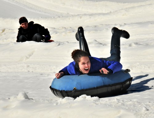 A girl joyfully sleds down a snowy hill on a blue tube, while another person sits in the background.