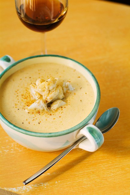 A bowl of creamy soup topped with crab meat, garnished with spices, next to a glass of wine on a wooden table.