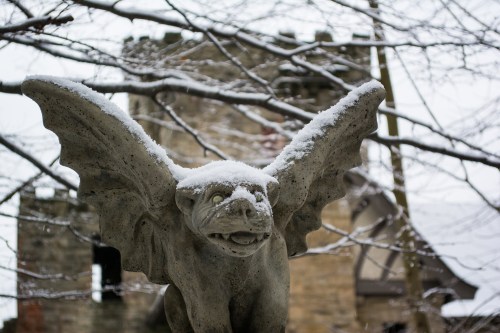 A snow-covered gargoyle with wings, set against a blurred stone castle in winter.