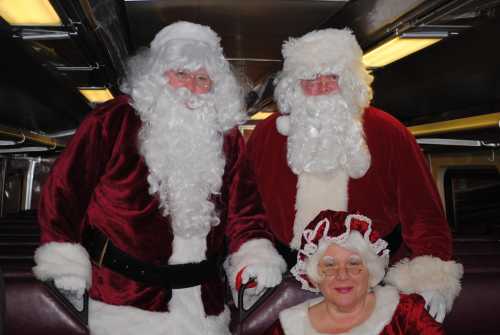 Two men in Santa suits and a woman in a festive outfit pose together on a train.