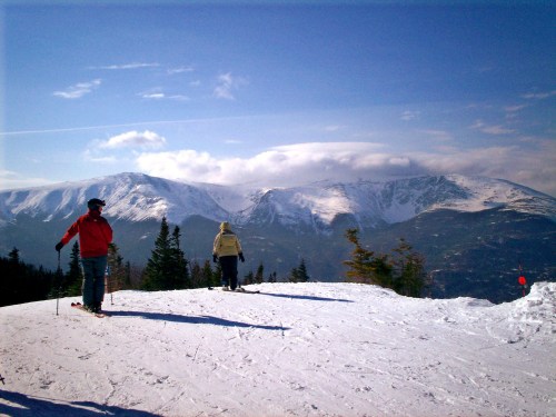 Two skiers on a snowy mountain slope, with majestic peaks and a clear blue sky in the background.