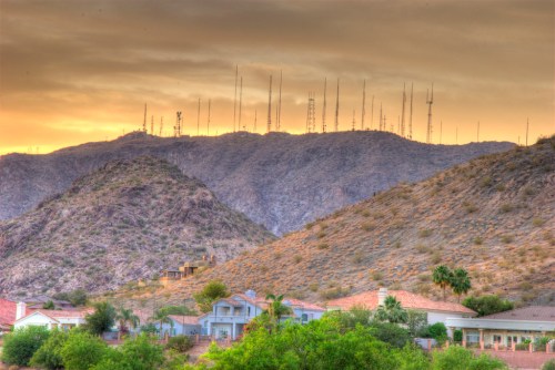 A mountainous landscape at sunset, featuring radio towers on the peak and homes in the foreground surrounded by greenery.