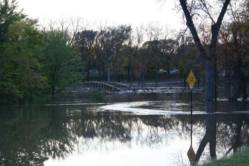 A flooded area with a submerged bridge and a warning sign, surrounded by trees and reflecting water.