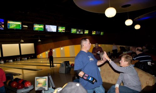 A man in a blue shirt celebrates while a woman playfully gestures at him in a bowling alley with lanes in the background.
