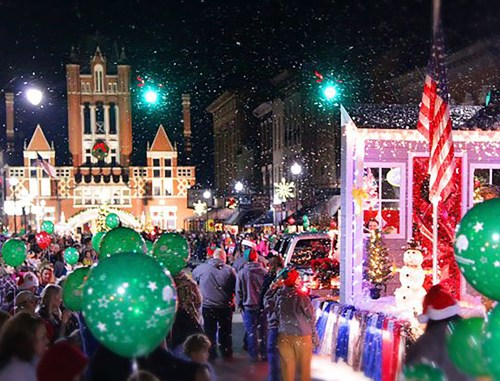 A festive night scene with a parade, decorated floats, and people holding green balloons in a snowy atmosphere.
