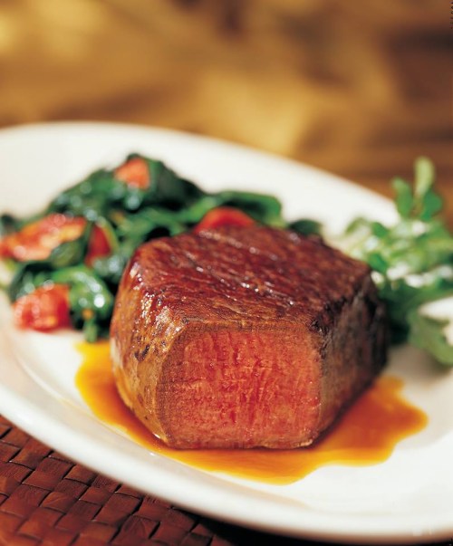A perfectly cooked steak on a plate, accompanied by sautéed greens and cherry tomatoes, drizzled with sauce.