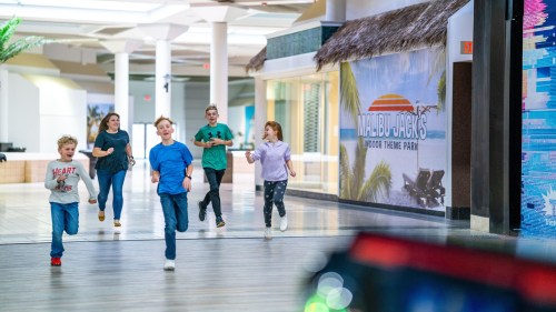 A group of children and adults run joyfully through a mall towards Malibu Jack's indoor theme park.