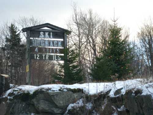 Sign for "Ski Back" with "12 Trails" displayed, surrounded by snow and trees, under a cloudy sky.