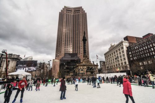 A bustling ice skating rink in an urban setting, with a tall building and cloudy sky in the background.