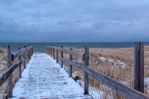 A wooden boardwalk leads to a snowy beach, with calm waters and overcast skies in the background.
