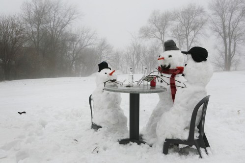 Three snowmen sitting at a table in a snowy landscape, wearing hats and scarves, with drinks in front of them.