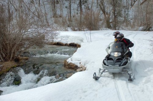 Two people on a snowmobile near a flowing stream in a snowy landscape surrounded by trees.