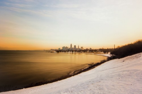 A serene winter landscape with snow-covered ground and a city skyline at sunset over a calm lake.