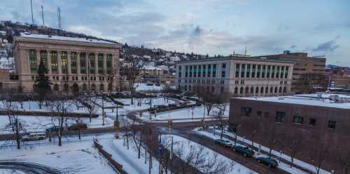 A snowy urban landscape featuring buildings, trees, and a winding road under a cloudy sky.