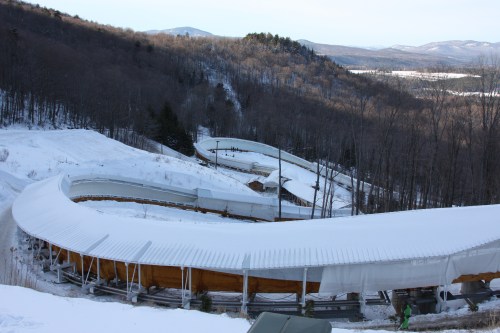 A snowy landscape featuring a winding bobsled track surrounded by trees and mountains in the background.