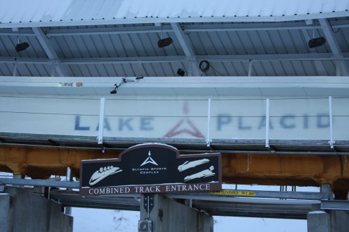 Sign for the Combined Track Entrance at Lake Placid, with a snowy roof overhead and a blurred background.
