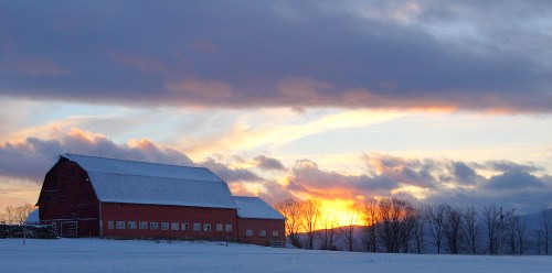 A red barn in a snowy landscape at sunset, with clouds and trees silhouetted against the colorful sky.