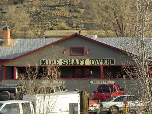A rustic tavern with a sign reading "Mine Shaft Tavern," surrounded by trees and parked vehicles.