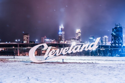 A snowy night view of a large "Cleveland" sign with the city skyline illuminated in the background.