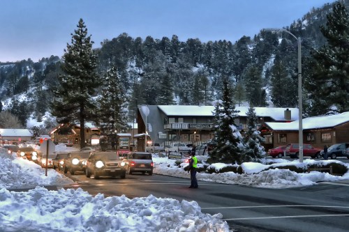 A snowy street scene with cars lined up, a traffic controller, and pine trees in the background at dusk.