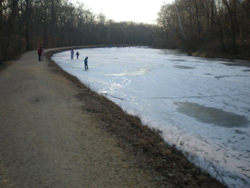 A frozen river with people skating and walking along a dirt path lined by trees in a winter landscape.