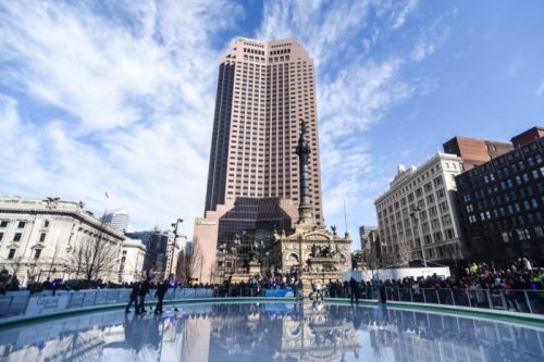 A bustling ice skating rink in a city square, surrounded by tall buildings and a clear blue sky.