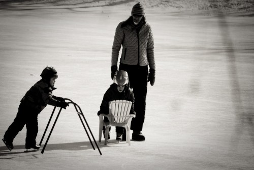 A woman watches two children on ice; one pushes a chair while the other sits, both wearing winter gear.