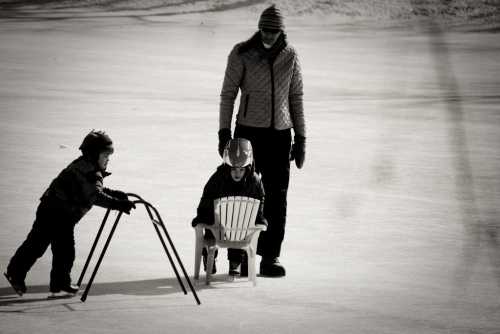 A woman watches two children on ice; one pushes a chair while the other sits, both wearing winter gear.