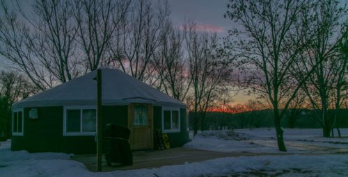 A yurt surrounded by snow-covered ground and bare trees, with a colorful sunset in the background.
