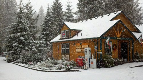A cozy yellow cabin surrounded by snow-covered trees, with snow falling gently and a gas pump in front.