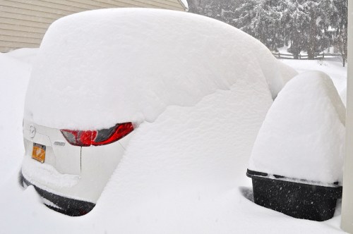 A car almost completely buried in snow, with only the rear and part of the roof visible, next to a snow-covered trash can.
