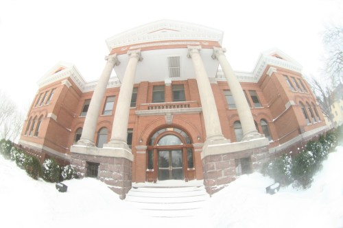 Historic brick building with large columns, partially covered in snow, under a cloudy sky.