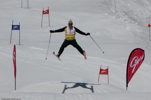 A skier jumps mid-air over a snow-covered slope, with flags and banners in the background.