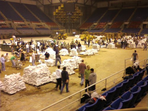 Volunteers packing and stacking bags in a large indoor arena, with machinery and empty seats in the background.
