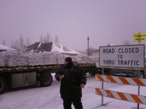 A person in winter gear stands near a sign that reads "Road Closed to Thru Traffic," with a truck loaded with sandbags behind.