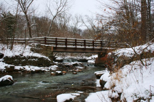 A snow-covered bridge spans a serene stream, surrounded by bare trees and rocky banks in a winter landscape.