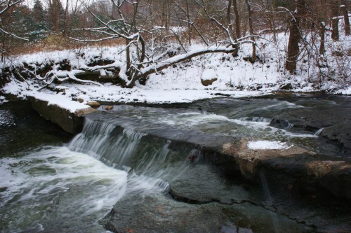 A serene winter scene featuring a small waterfall flowing over rocks, surrounded by snow-covered trees and a tranquil stream.