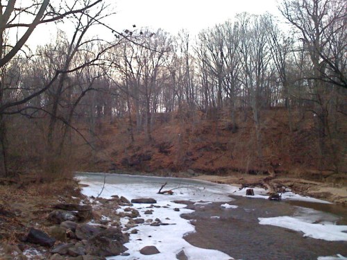 A serene winter landscape featuring a river with ice, surrounded by bare trees and a rocky shoreline.
