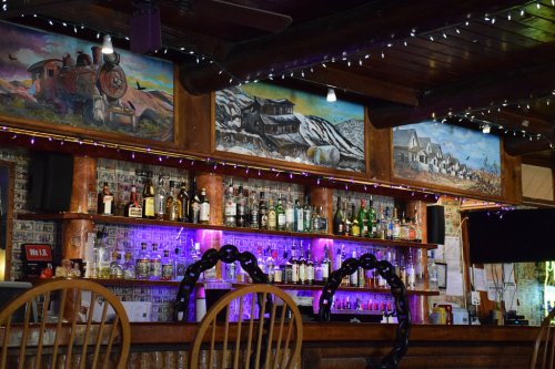 A rustic bar with a wooden counter, colorful liquor bottles, and murals of trains and landscapes above.