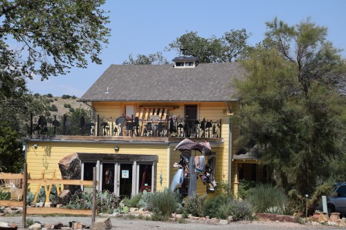 A two-story yellow house with a balcony, surrounded by trees and desert plants, featuring outdoor seating.