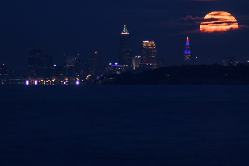 A city skyline at night with a large, glowing moon rising above, casting light over the water.