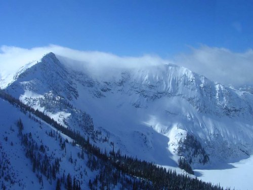 Snow-covered mountains under a clear blue sky, with clouds gently resting on the peaks. Pine trees line the lower slopes.