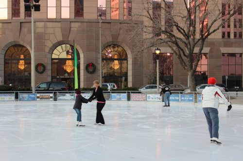 People ice skating on a rink with festive decorations and buildings in the background during winter.