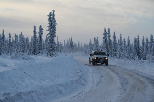 A vehicle drives along a snowy, winding road surrounded by tall, snow-covered trees under a cloudy sky.