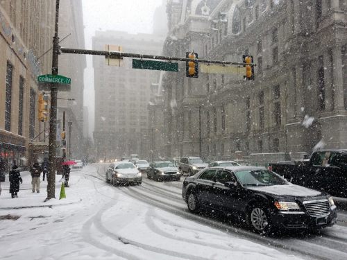 A snowy city street with cars navigating through heavy snowfall and pedestrians walking along the sidewalk.