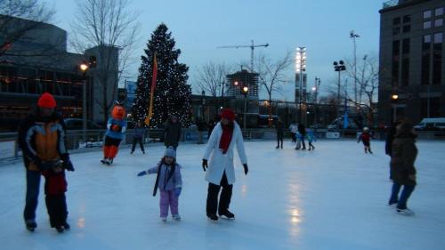 A winter scene at an ice rink with skaters, a Christmas tree, and festive decorations in the background.