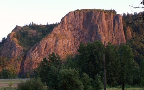 A large, rugged rock formation bathed in warm light, surrounded by lush green trees and a clear sky.