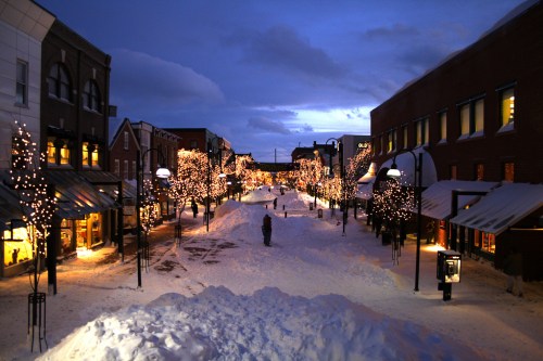 A snowy street lined with trees adorned with lights, illuminated shops, and a twilight sky.
