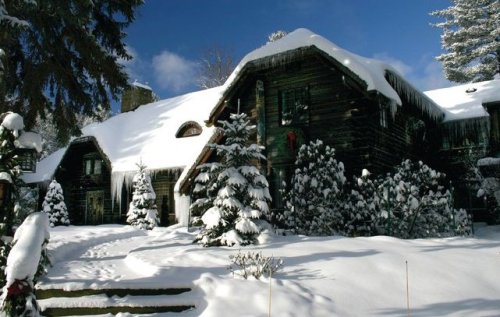 A snow-covered cabin surrounded by evergreen trees, with icicles hanging from the roof under a bright blue sky.