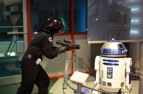 A person in a black costume aims a blaster at a blue and white droid in a futuristic setting.